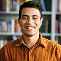 Man with dark hair, wearing burnt orange shirt, smiling into camera with bookshelves in the background